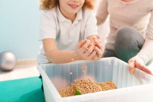 A child smiles while lifting a hand full of chickpeas from a plastic container. This could represent the support for child counseling in Boulder, CO that a play therapist for children in Boulder, CO can offer. Learn more about parent coaching in Boulder, CO and other services today. 