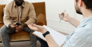 A close up of a man with a clipboard gesturing with his hands while sitting across from a black man. This could represent the support a depression therapist in Boulder, CO can offer. Overcome signs of depression in Boulder, CO by searching for depression treatment in Boulder, CO.