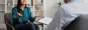 A smiling woman gestures with her hands and sitting across from a person with a clipboard. This could represent the support an anxiety therapist in Boulder, CO can offer. Learn more about overcoming symptoms of postpartum anxiety in Boulder, CO and other services. Search for anxiety treatment in Boulder, CO for more info